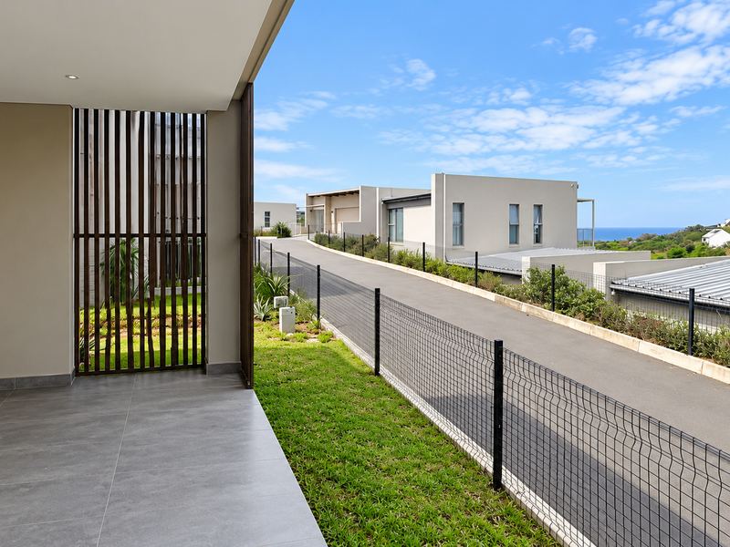 A covered patio area with modern grey floor tiles, recessed lighting, and a timber privacy screen, overlooking the estate driveway and coastal landscape.