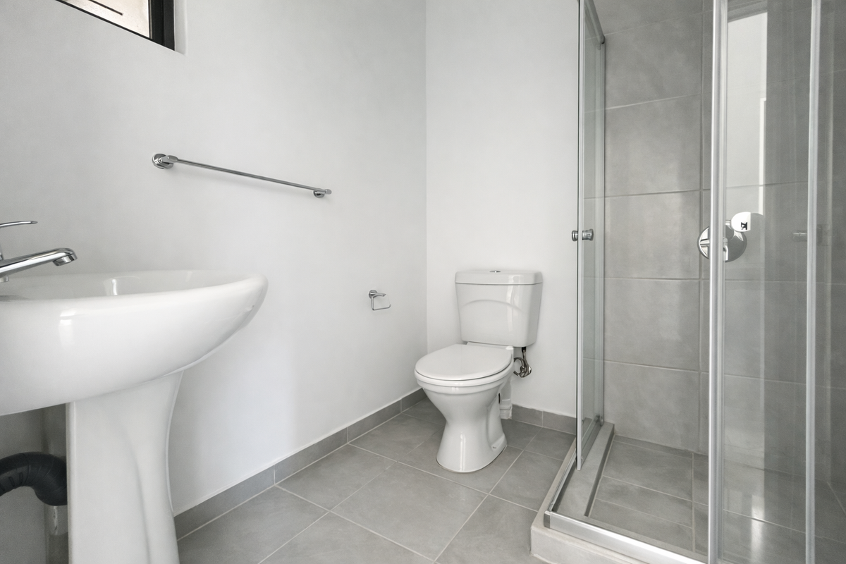 A clean, modern bathroom featuring a pedestal washbasin, a standard toilet, and a glass-enclosed shower with neutral grey floor and wall tiling.