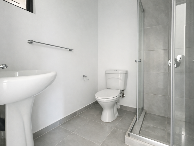 A clean, modern bathroom featuring a pedestal washbasin, a standard toilet, and a glass-enclosed shower with neutral grey floor and wall tiling.