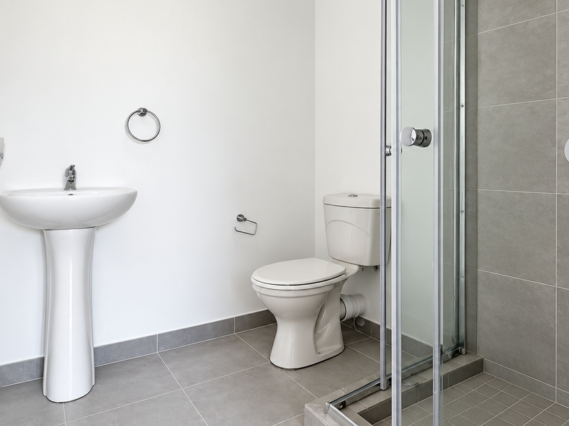 A clean, modern bathroom featuring a pedestal sink, a standard toilet, and a glass-enclosed shower with grey tiled finishes.