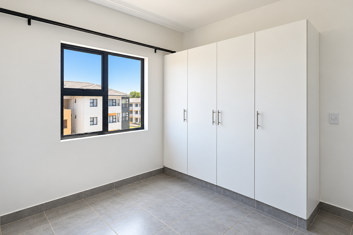 A bright, empty bedroom featuring modern tiled flooring, built-in white cupboards, and an aluminium-framed window with a curtain rod.