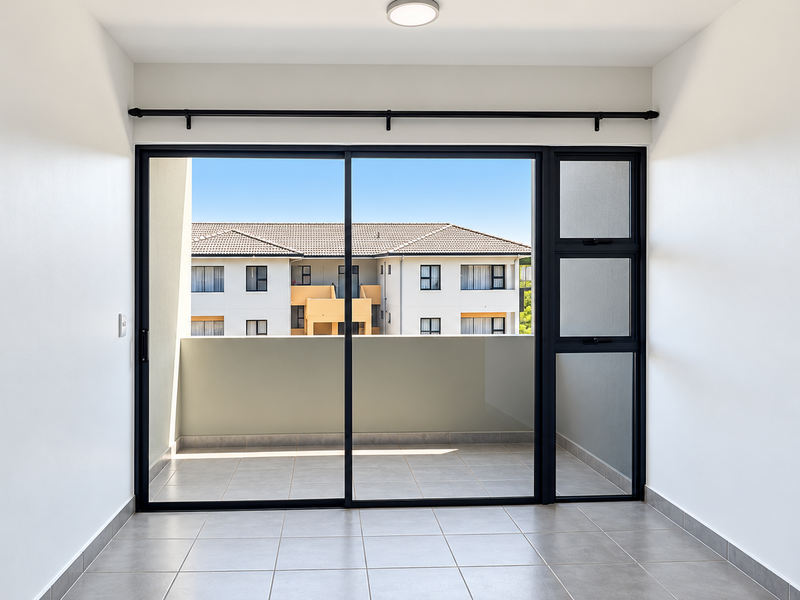 A bright, empty balcony area featuring tiled flooring and a large sliding glass door, offering a clear view of the surrounding apartment complex.