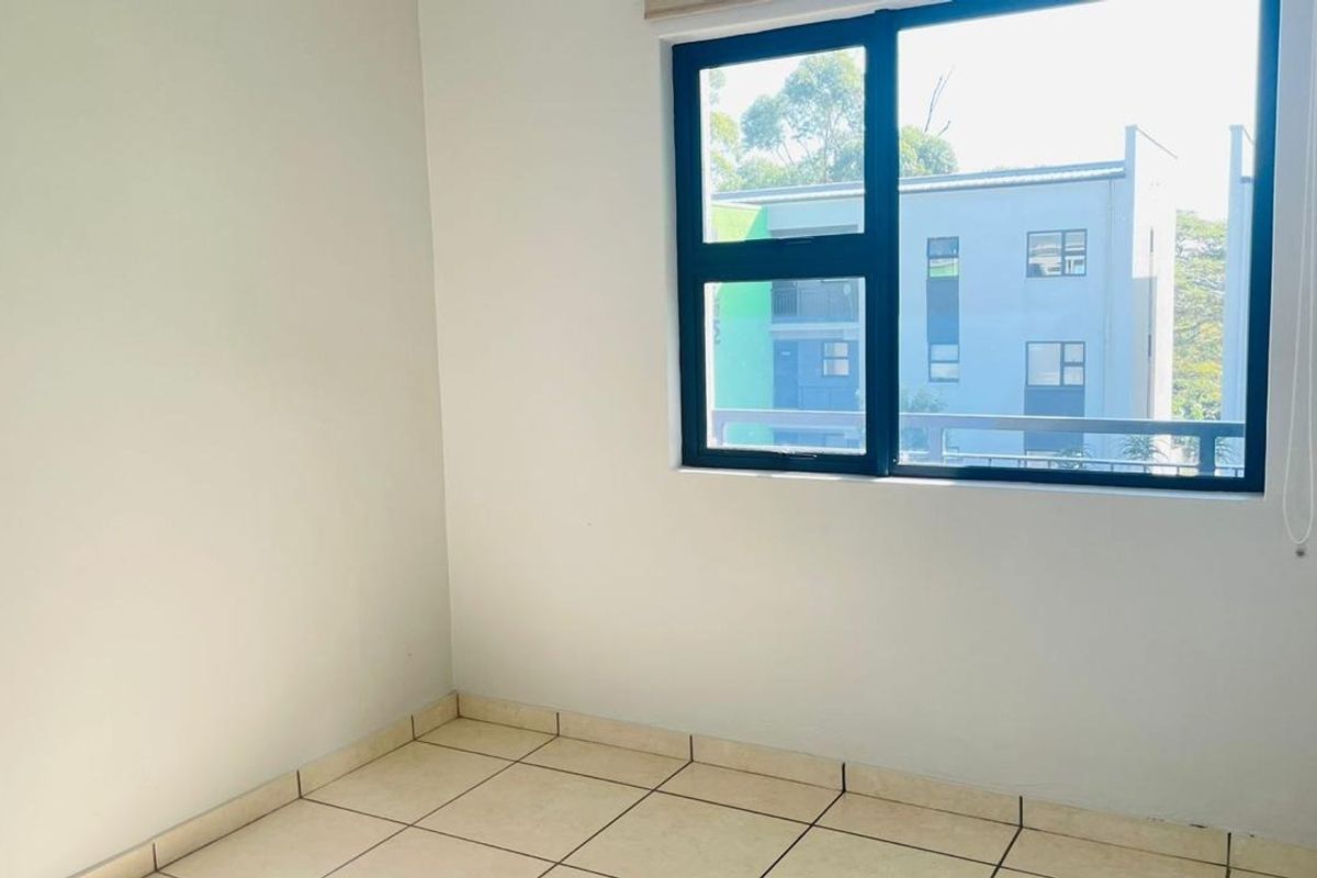 A bright, empty bedroom featuring neutral tiled flooring and a large aluminium-framed window providing natural light and an exterior view.
