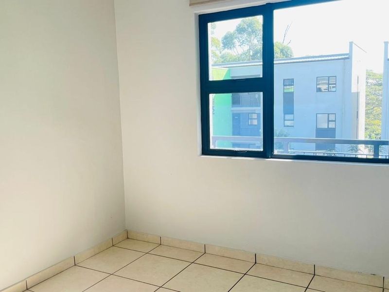 A bright, empty bedroom featuring neutral tiled flooring and a large aluminium-framed window providing natural light and an exterior view.