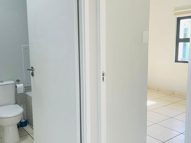A view from a hallway looking into a bathroom with a toilet and an adjacent bedroom with tiled flooring and neutral walls.