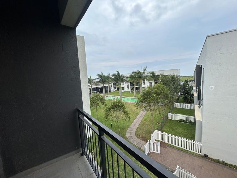 A view from a private balcony overlooking a landscaped communal garden, paved pathways, and a swimming pool area within the apartment complex.