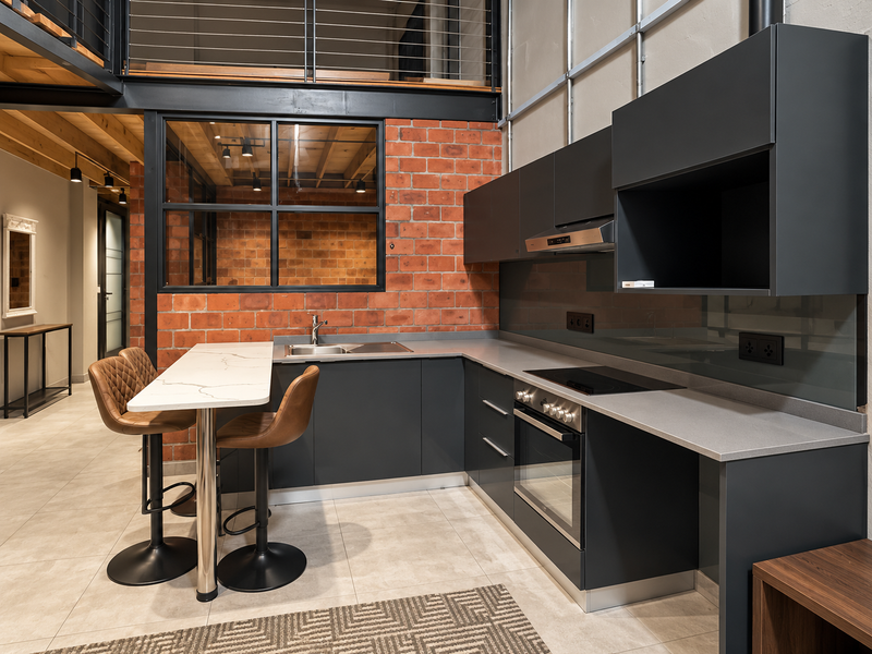 Modern industrial-style kitchen featuring dark cabinetry, stone countertops, integrated appliances, and an exposed brick wall with a mezzanine level above.