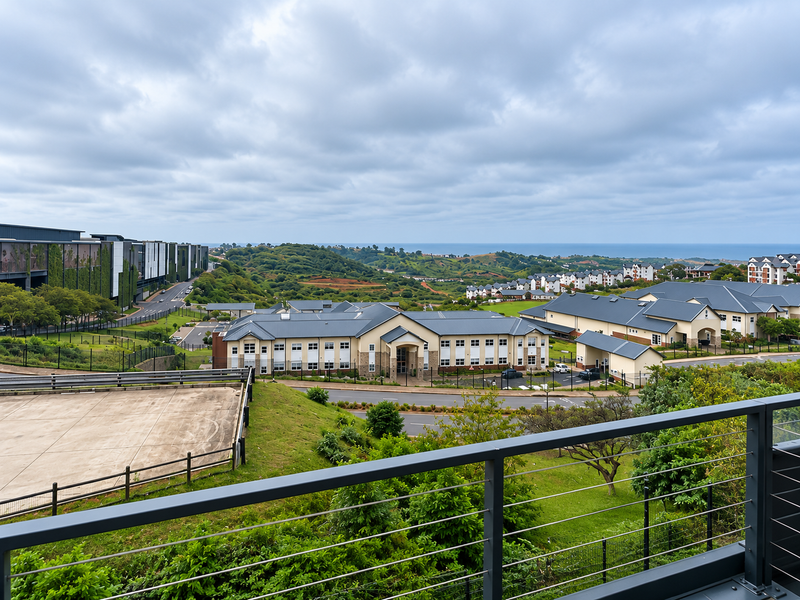 An elevated view overlooking a residential estate, landscaped grounds, and distant coastal scenery, captured from a modern balcony with a metal cable railing.
