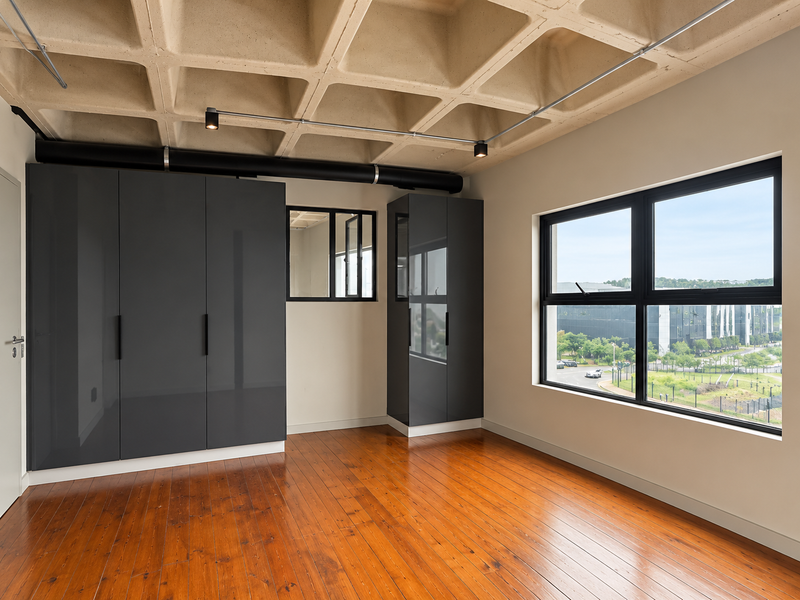 A modern, empty bedroom featuring a unique exposed concrete waffle ceiling, polished hardwood floors, built-in dark grey cabinetry, and large windows with exterior views.