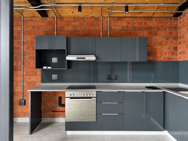 Modern industrial-style kitchen featuring grey cabinetry, stone countertops, built-in oven, electric hob, and exposed brick walls with timber ceiling beams.
