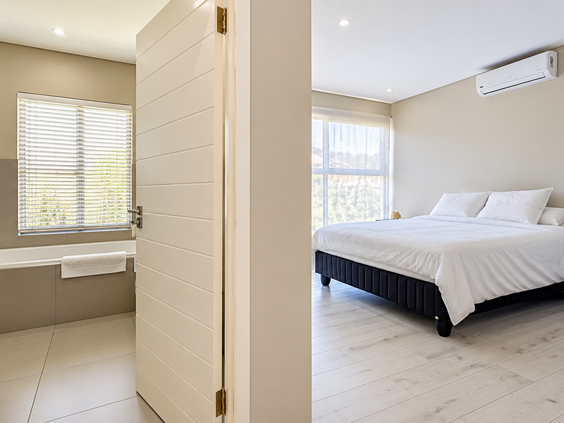 A modern bedroom featuring light wood-look flooring, a wall-mounted air conditioner, and an adjoining en-suite bathroom with a bathtub and glass shower.