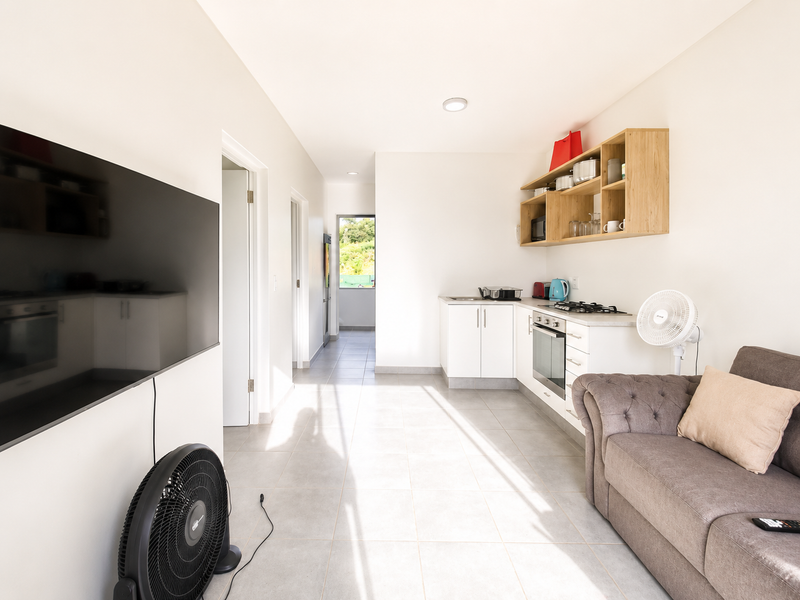 An open-plan living and kitchen area featuring tiled flooring, modern white cabinetry, built-in cooking appliances, and open wooden shelving.
