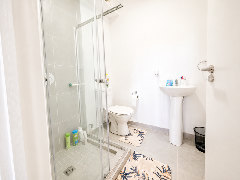 A clean and functional bathroom featuring a pedestal basin, a toilet, and a glass-enclosed shower with neutral grey floor and wall tiling.