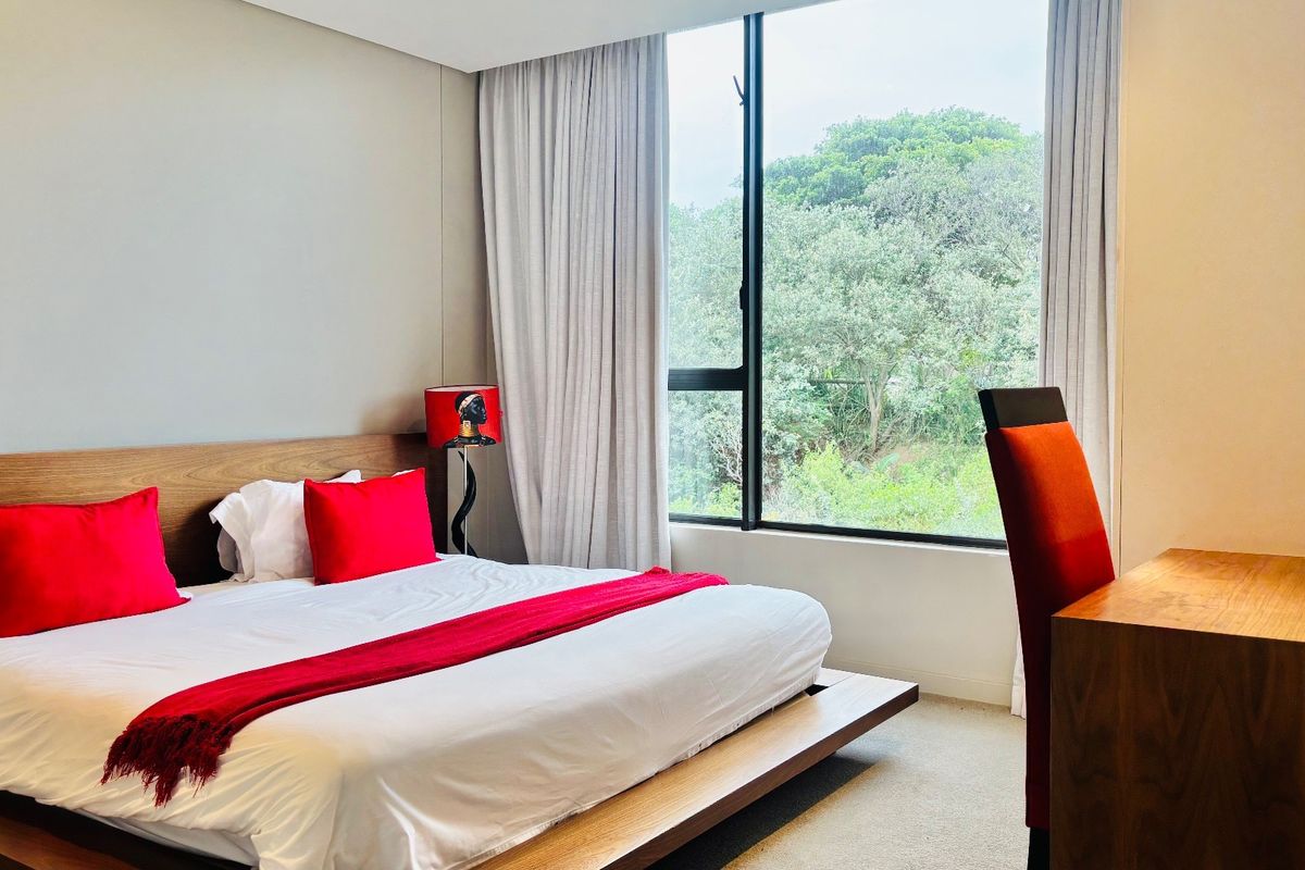 A well-maintained bedroom featuring large windows with curtain tracks, carpeted flooring, and a pleasant view of the surrounding greenery.