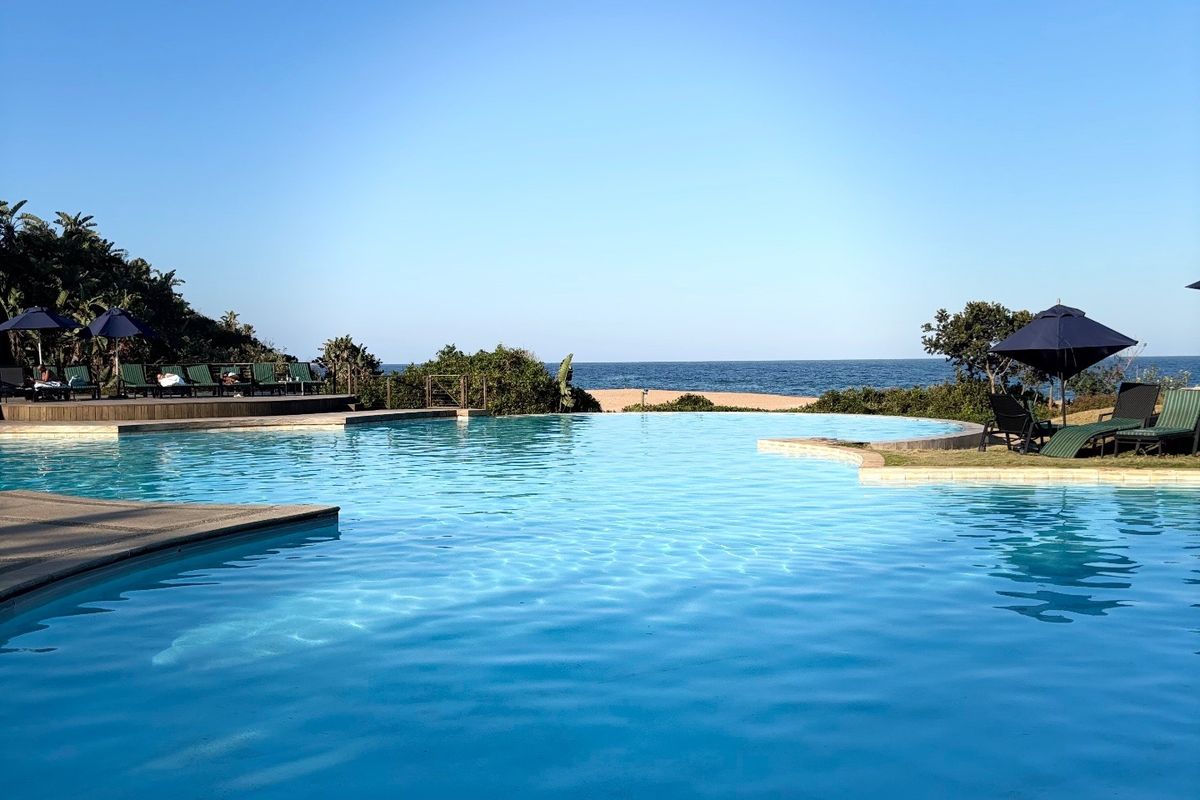 A pristine infinity-style swimming pool featuring timber decking and landscaped surroundings, offering direct views of the ocean and beach.