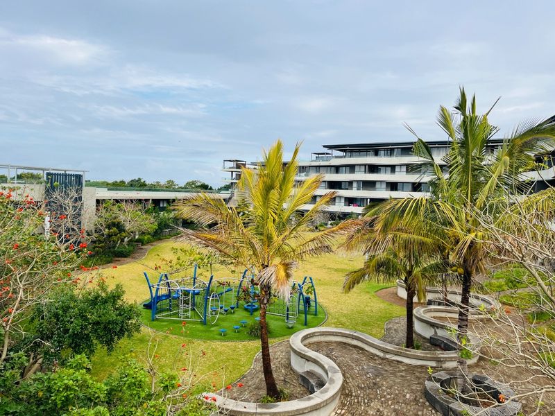 A well-maintained communal garden area featuring landscaped lawns, mature palm trees, paved walkways with curved concrete retaining walls, and a children's playground.