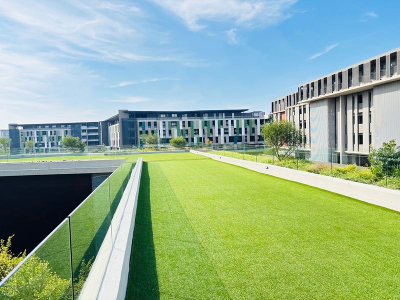 A well-maintained rooftop garden area featuring artificial turf and modern glass balustrades, overlooking the surrounding apartment complex.
