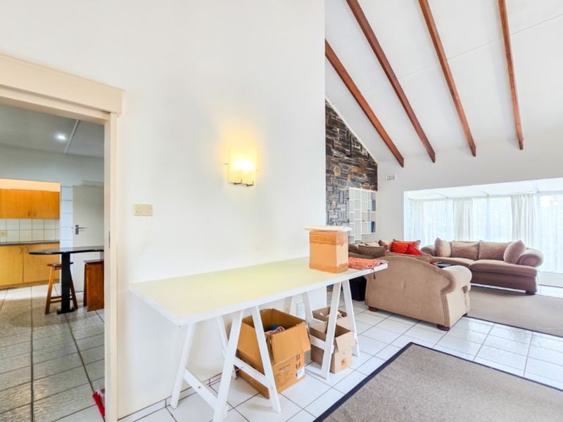 Open plan living area featuring exposed timber ceiling beams, a stone feature wall, and tiled flooring, with an adjacent kitchen visible through the doorway.