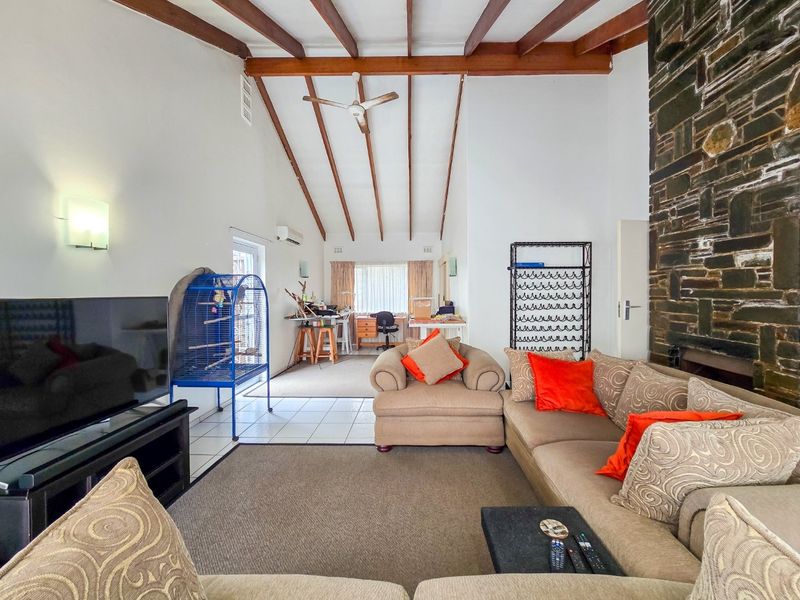 Spacious living room featuring high vaulted ceilings with exposed timber beams, a prominent stone fireplace, and tiled flooring with an adjoining workspace area.