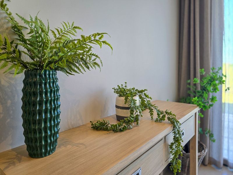 A close-up view of a wooden console table with decorative plants, positioned against a neutral wall with curtains in the background.