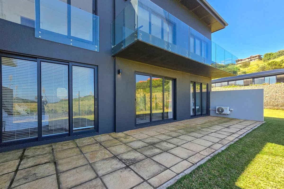 Rear exterior view of a modern apartment featuring a paved patio, glass balustrade balcony, and sliding door access to the living areas.