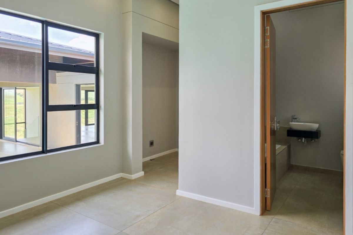 A modern, empty bedroom featuring large aluminium windows, neutral tiled flooring, and an adjoining en-suite bathroom with a wall-mounted basin and built-in bathtub.