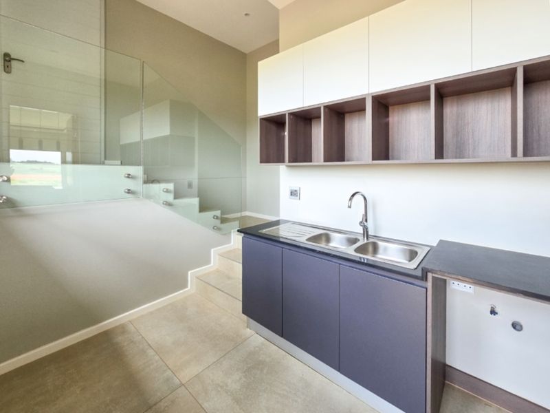 A modern, minimalist scullery area featuring sleek cabinetry, a double sink, and a glass balustrade staircase in a clean, contemporary setting.
