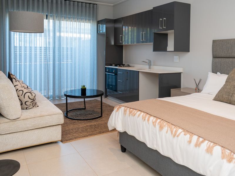 Modern open-plan studio apartment featuring a compact kitchenette with built-in cabinetry, integrated appliances, and direct access to a balcony through sliding glass doors.
