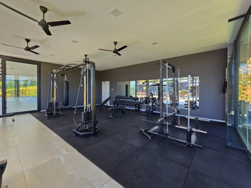 A modern, well-equipped home gym featuring rubber flooring, large wall mirrors, recessed lighting, ceiling fans, and expansive floor-to-ceiling glass walls.
