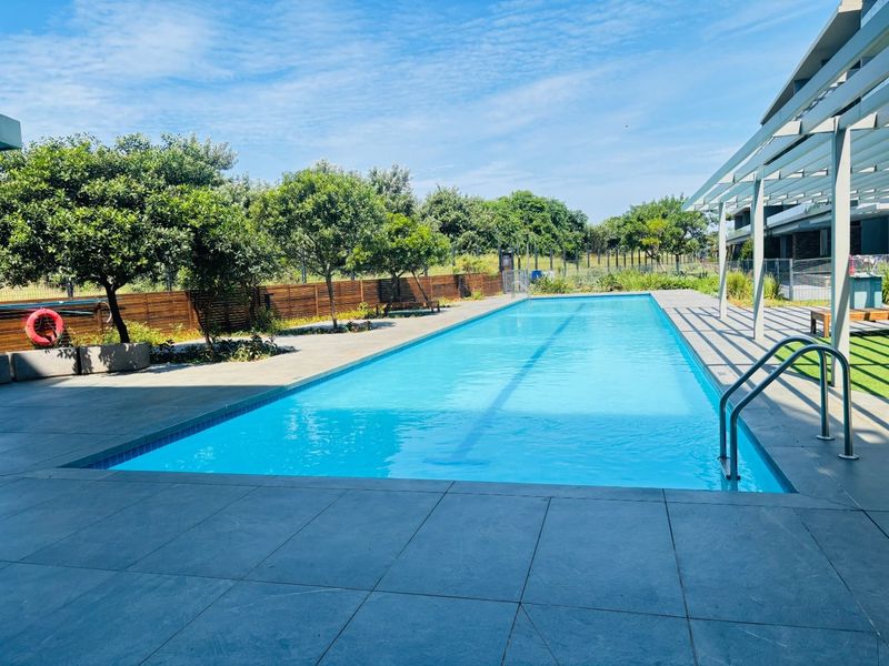 A well-maintained outdoor swimming pool area featuring a long lap pool, paved decking, and a modern pergola structure.