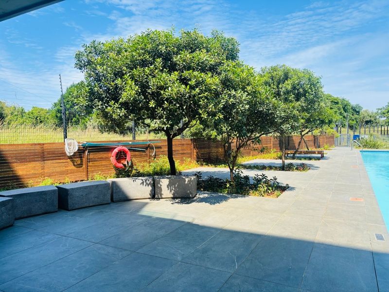 Well-maintained outdoor pool area featuring a tiled deck, concrete bench seating, mature trees, and secure perimeter fencing.