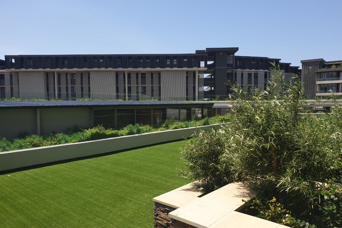 A well-maintained outdoor terrace featuring artificial turf, stone-clad retaining walls with sandstone coping, and a glass balustrade overlooking the apartment complex.