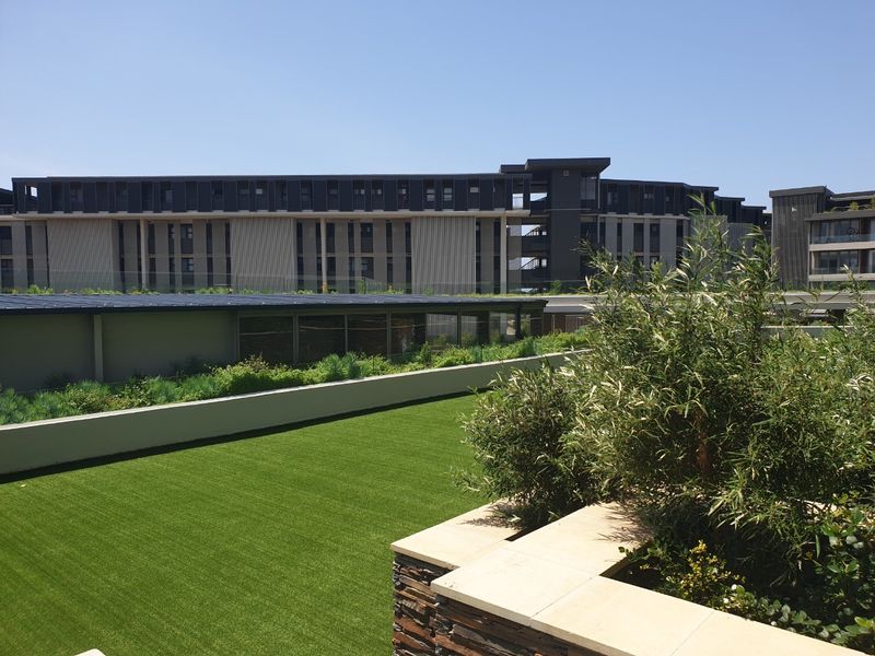 A well-maintained outdoor terrace featuring artificial turf, stone-clad retaining walls with sandstone coping, and a glass balustrade overlooking the apartment complex.