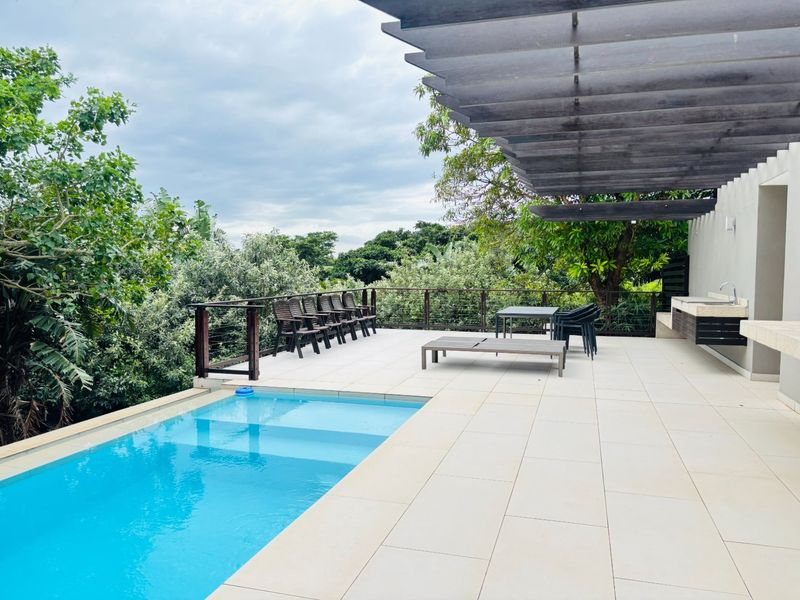 Spacious outdoor patio area featuring a modern swimming pool, a wooden pergola, and a built-in counter, set against a lush garden backdrop.