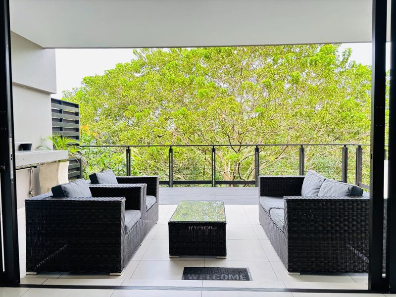 A covered patio featuring tiled flooring, a built-in braai, and a metal balustrade overlooking lush greenery.