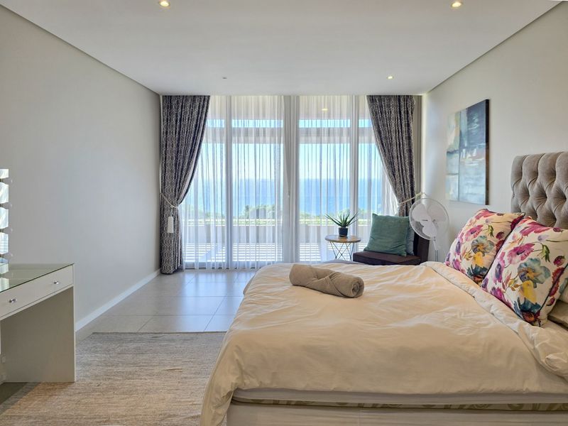 Spacious bedroom featuring neutral tones, tiled flooring, and floor-to-ceiling glass doors that open to a balcony with ocean views.