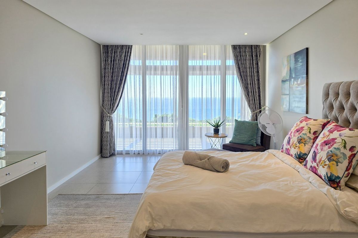 Spacious bedroom featuring neutral tones, tiled flooring, and floor-to-ceiling glass doors that open to a balcony with ocean views.