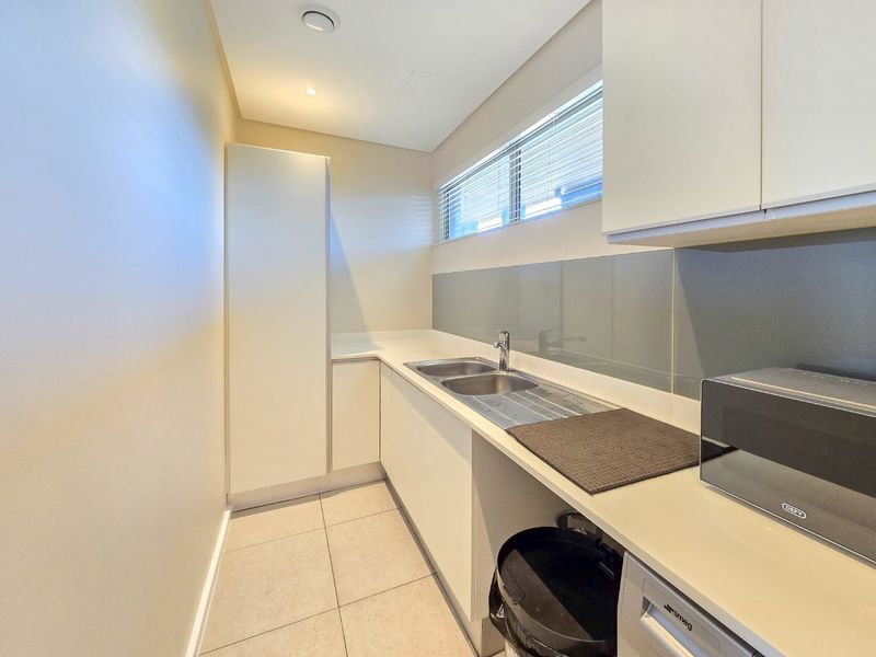 A clean and functional scullery featuring modern white cabinetry, stone countertops, a double stainless steel sink, and a sleek glass splashback.