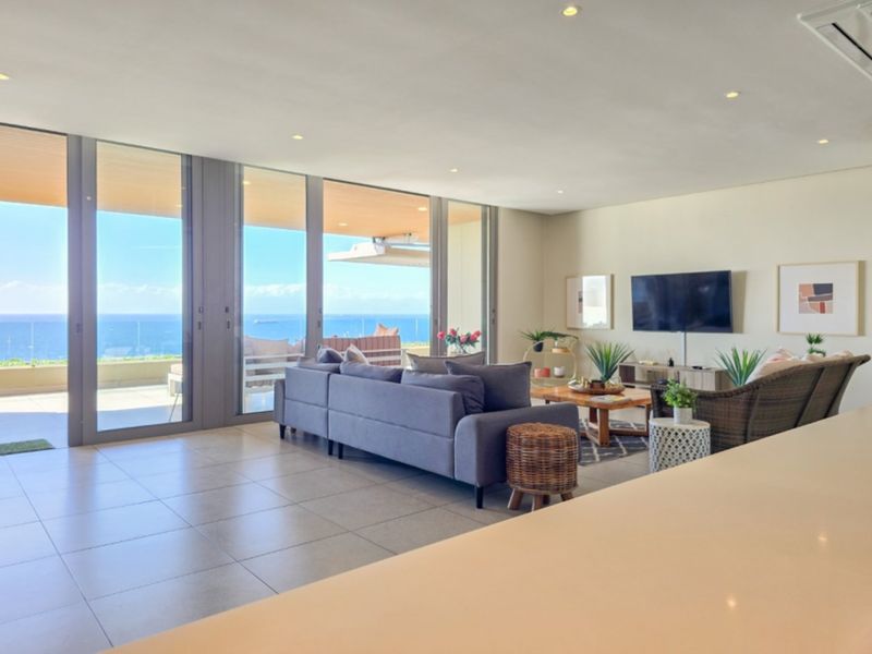 Spacious open-plan living area featuring large format floor tiles, recessed lighting, and expansive floor-to-ceiling glass doors opening to a scenic ocean view.