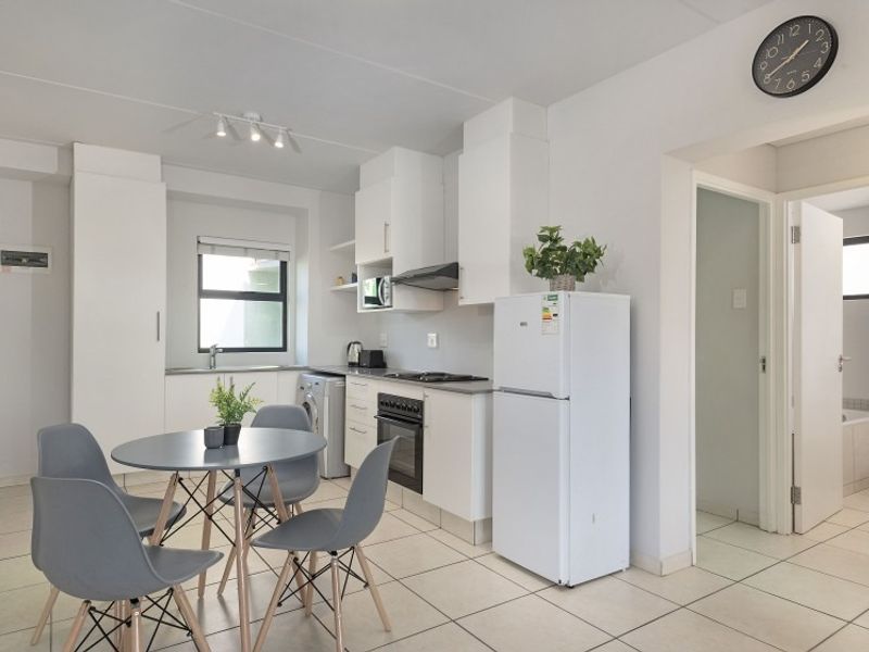 A bright open-plan kitchen and dining area featuring white cabinetry, tiled floors, and integrated appliances, with direct access to an adjacent bathroom.