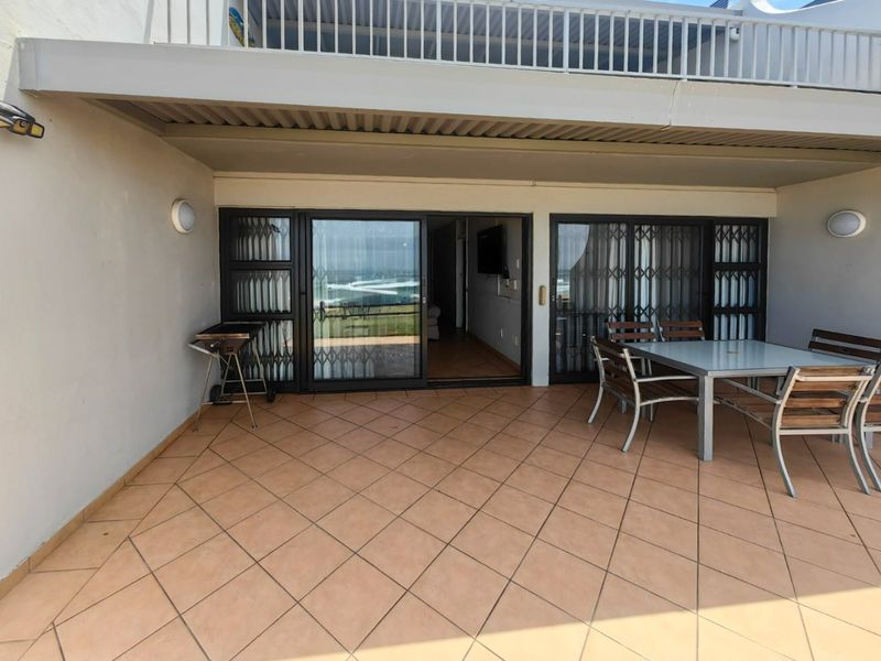 A covered patio area featuring tiled flooring, sliding glass doors with security gates, and wall-mounted lighting, providing a functional outdoor living space.