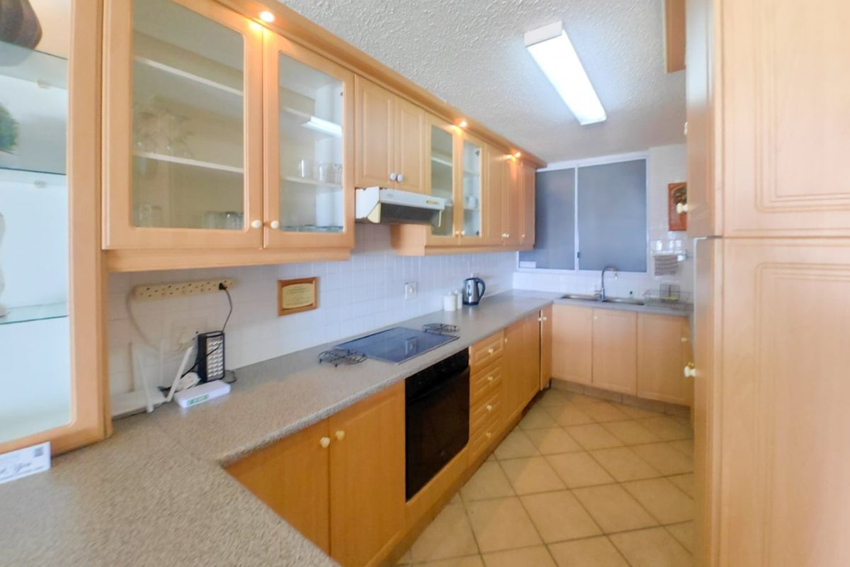 A functional galley-style kitchen featuring light wood cabinetry, laminate countertops, integrated oven and hob, and tiled flooring.