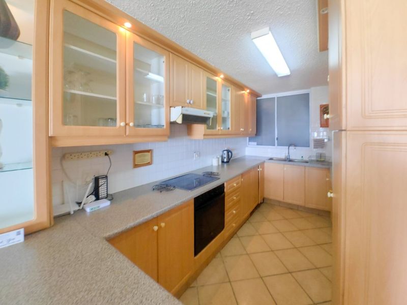 A functional galley-style kitchen featuring light wood cabinetry, laminate countertops, integrated oven and hob, and tiled flooring.