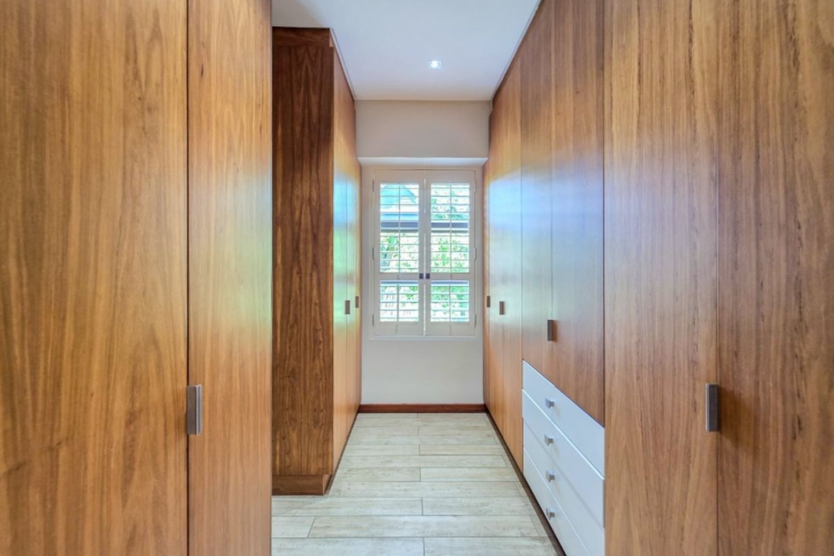 A well-appointed walk-in dressing area featuring extensive floor-to-ceiling timber veneer built-in cabinetry, tiled flooring, and plantation shutters.
