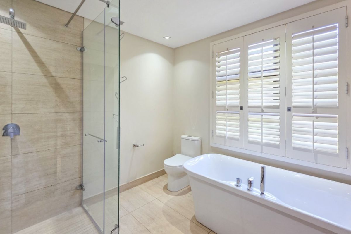 Modern bathroom featuring a freestanding bathtub, a spacious glass-enclosed rain shower, and elegant plantation shutters, finished with neutral floor-to-ceiling tiling.