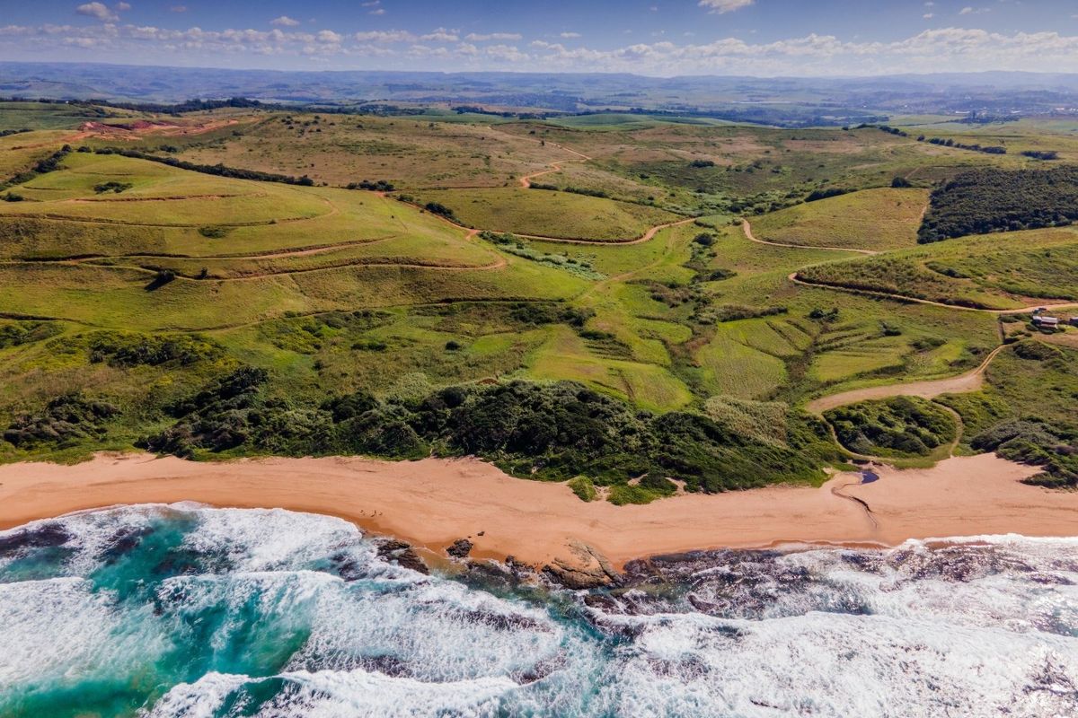 An aerial view of expansive vacant coastal land featuring rolling green hills, natural vegetation, and direct access to a sandy beach and ocean.