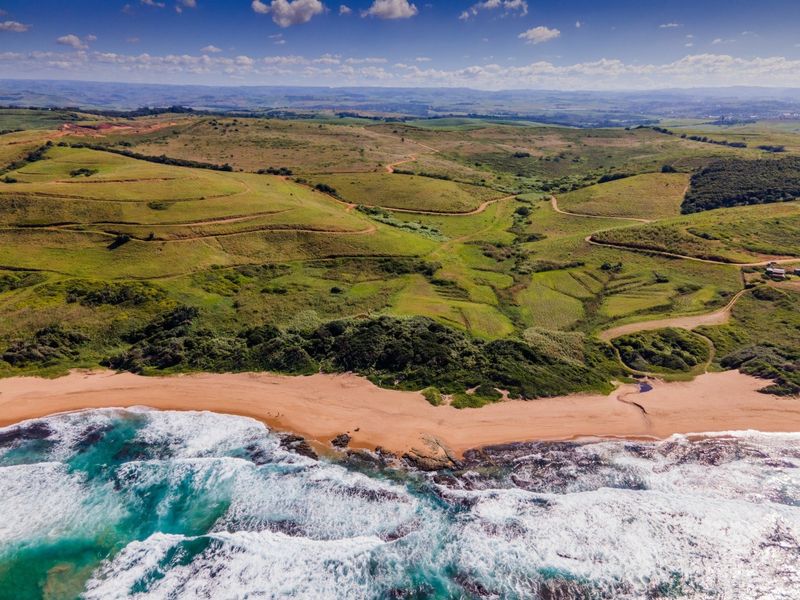 An aerial view of expansive vacant coastal land featuring rolling green hills, natural vegetation, and direct access to a sandy beach and ocean.