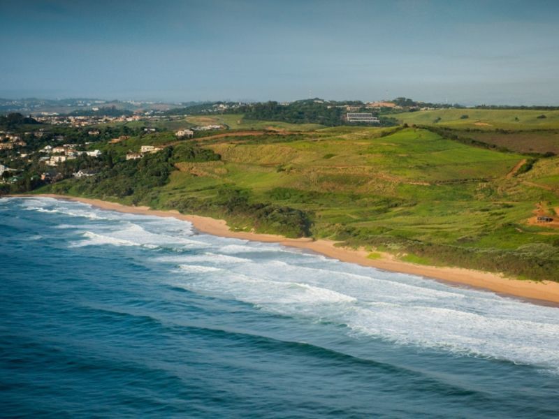 An aerial view of the coastline featuring sandy beaches, rolling green hills, and residential developments in the distance.
