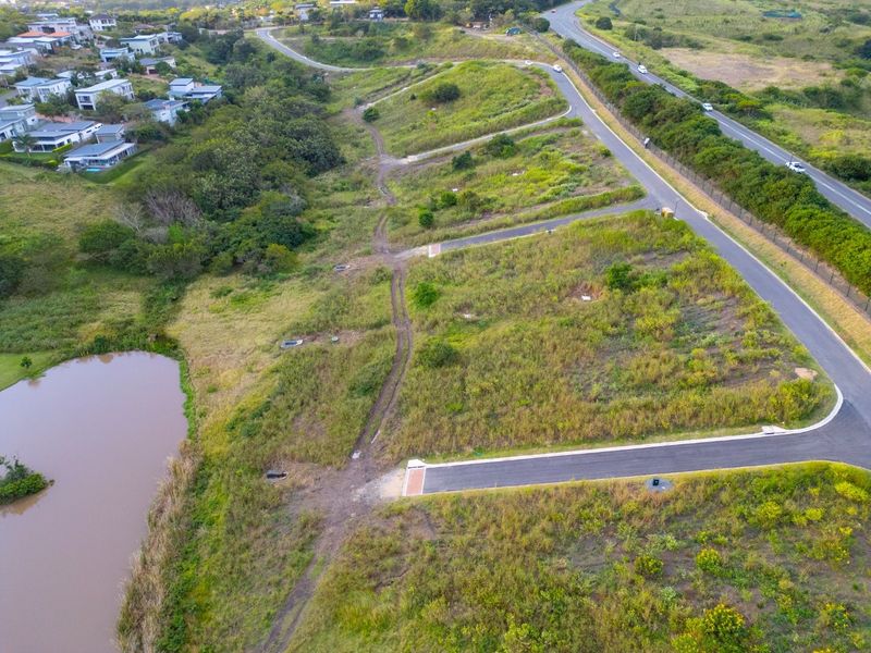 Aerial view of a vacant residential land parcel featuring established road access, drainage infrastructure, and proximity to a natural pond and surrounding residential development.