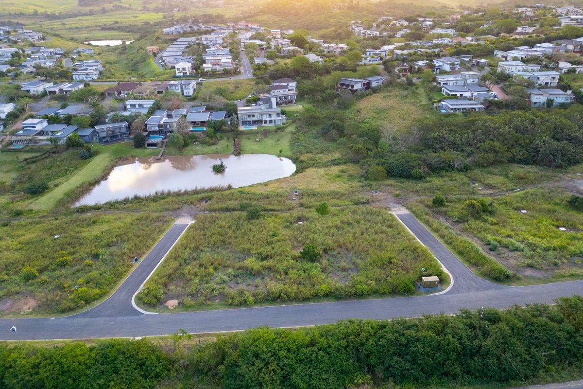 Aerial view of a vacant residential plot featuring paved road access and proximity to a scenic estate pond and surrounding neighborhood.